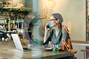 Woman at cafe making a phone call and using laptop computer. Beautiful young woman sitting at coffee shop working on laptop and