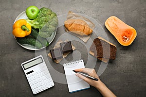 Woman calculating calories at dark grey table with food products, top view. Weight loss concept