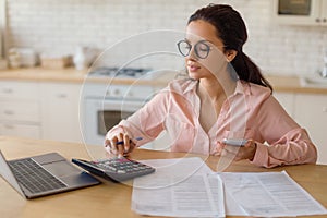 Woman calculating with smartphone and calculator at kitchen table