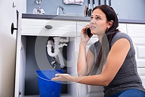 Woman Calling Plumber While Collecting Water Leaking From Sink