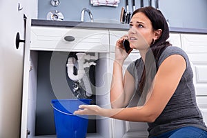 Woman Calling Plumber While Collecting Water Leaking From Sink photo