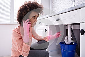 Woman Calling Plumber In Front Of Leaking From Sink Pipe