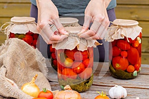 Woman cans pickled tomatoes in glass jars on an old wooden table. Summer harvest.