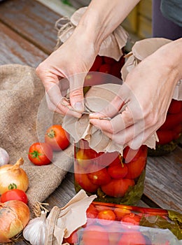 Woman cans pickled tomatoes in glass jars on an old wooden table. Summer harvest.