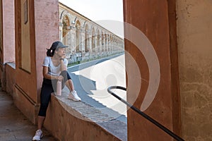 Woman in cap and sneakers holding thermos and sitting in window of portico with chin on her hand