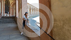 Woman in cap and sneakers holding thermos and sitting in window of portico with chin on her hand