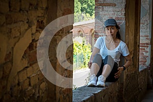 Woman in cap and sneakers sitting in window of portico looking at camera