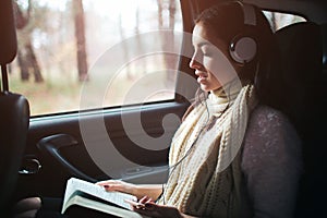 Woman in the car, autumn concept. Smiling pretty girl listening to music with headphones and reading a book moving in