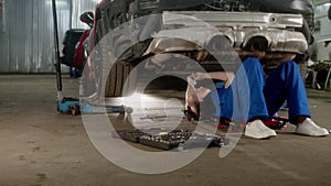 Woman car service worker lies under car and repairs it. female mechanic in blue overall with ratchet wrench maintains