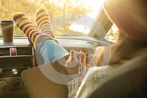 Woman in a car in warm socks is writing notes and plans in a notebook. Cozy autumn weekend trip. Freedom of travel