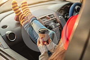 Woman in a car in warm socks yellow is holding a Cup of coffee in hands at sunset. Cozy autumn weekend trip