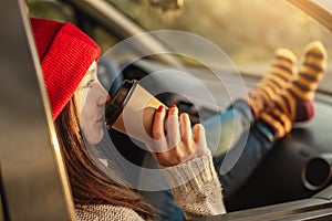 Woman in a car in warm socks yellow is holding a Cup of coffee in hands at sunset. Cozy autumn weekend trip