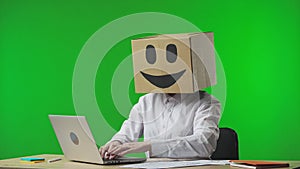 Woman with cardboard box on her head on green studio background. Worker with sad emoji is thinking about something, an
