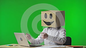 Woman in cardboard box with smiling emoji on her head on studio green background. The female worker is typing on a