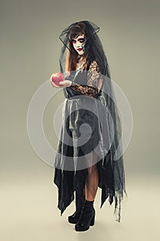 Woman in a carnival costume of a witch or a dead bride holding an apple in her hands, gothic woman in witch costume on gray