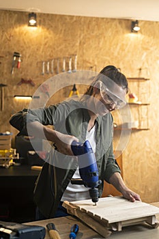 Woman carpenter with safety goggles working with drill in workshop