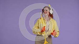 Woman in casual clothing and rubber gloves in headphones dancing with broom, playing on it like a guitar, singing and