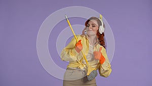 Woman in casual clothing and rubber gloves in headphones playing on broom like a guitar and smiling.  on purple