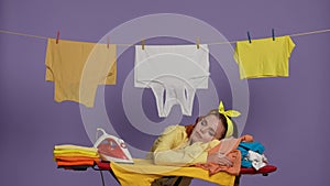 Woman in casual clothing sleeping on pile of shirts laying on the ironing board, laundry hanging behind her. Isolated on
