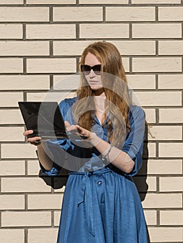 Woman in casual wear leans against white brick wall. Red hair girl is standing outside an office, holding a tablet