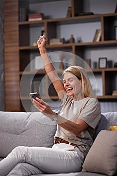 Woman celebrating victory by showing yes gesture on sofa with smartphone. Overjoyed to win, read the good news on the