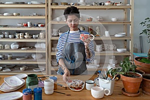 Woman ceramist having morning snack in ceramics workshop, drinking tea with sweet delicious bun