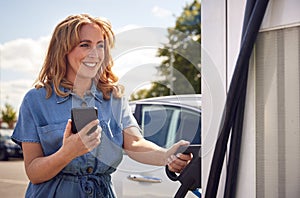 Woman Charging Electric Car With Cable Using App On Phone To Monitor Battery Level