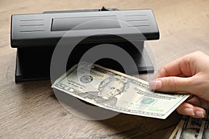 Woman checking dollar banknote with currency detector at wooden table, closeup. Money examination device