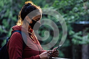 Woman checking mobile and wearing face mask while walking on nature environment. Covid-19 outbreak