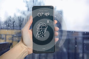 Woman checking weather using app on smartphone near wet window, closeup. Data and illustration of cloud with lightning and rain on