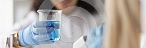 Woman chemist holding glass beaker with blue liquid in front of microscope in laboratory