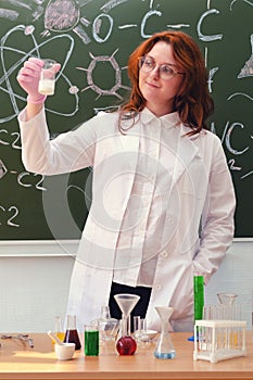 A woman chemistry teacher stands with white powder in a test tube near a painted blackboard. Teaching chemistry in a school class