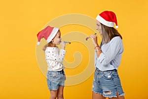 Woman child baby girl in Christmas Santa hat blow in pipe. Mommy little kid daughter isolated on yellow background