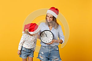 Woman child baby girl in Christmas Santa hat hold clock. Mommy little kid daughter isolated on yellow background studio