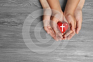 Woman and child holding heart with cross symbol on grey wooden background, top view. Christian religion