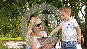 Woman and child using tablet computer. Slow motion. Young mother and little son spending day in the park.