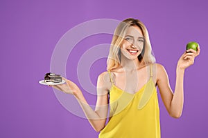 Woman choosing between cake and healthy apple on violet background