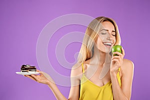 Woman choosing between cake and healthy apple on violet background