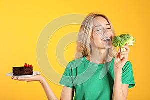 Woman choosing between cake and healthy broccoli on yellow background