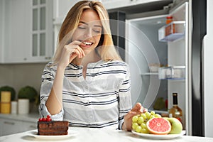 Woman choosing between cake and healthy fruits at table in kitchen