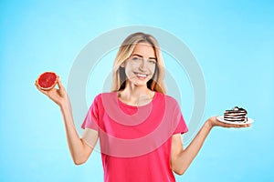 Woman choosing between cake and healthy grapefruit on light blue background