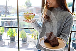 A woman choosing food to eat between a plate of brownie cake and a bowl of vegetables salad