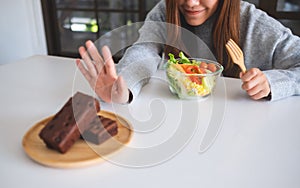 A woman choosing to eat vegetables salad and making hand sign to refuse a brownie cake