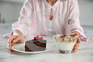 Woman choosing between yogurt with granola and cake at white table in kitchen, closeup