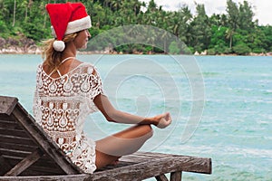 Woman in christmas hat meditating in a yoga pose at thel beach