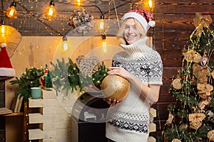 Woman in a Christmas room with gold bauble. The girl sits near Christmas tree , gifts and fireplace in home clothes.