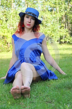 Woman in a classic blue short dress and hat sits stretching her legs forward on the green lawn. red hair and earrings