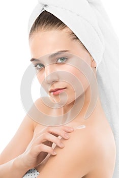 Woman with clean face and towel on her head applying moisturizer cream at shoulders. Isolated.