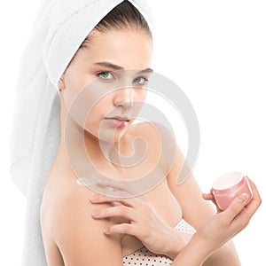 Woman with clean face and towel on her head applying moisturizer cream at shoulders. Isolated.