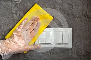 Woman clean light switch with cloth on gray concrete wall. Hand in glove disinfecting surfaces by yellow rag. The new normal Covid